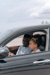 Father guides son during a driving lesson inside a black sedan on a bright day.